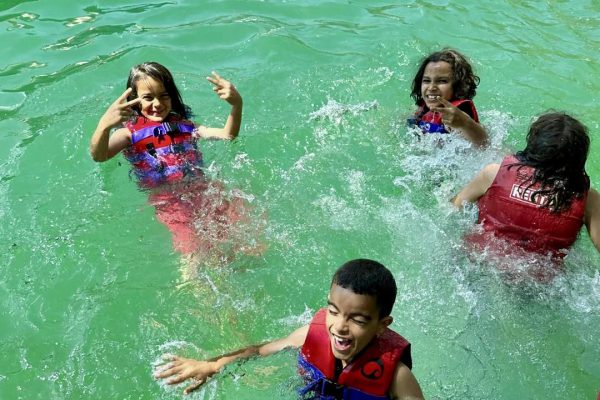 Groupe d'enfants souriants portant des gilets de sauvetage rouges, jouant dans l'eau verte d'un lac ou d'une piscine extérieure. Ils s'éclaboussent et s'amusent, profitant d'une activité aquatique par une journée ensoleillée